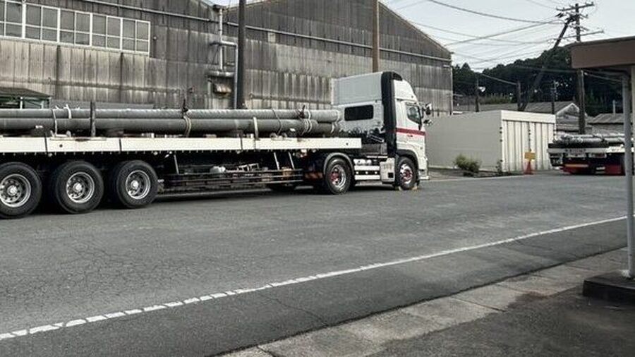工場前の道路を走る、電柱を積んだ長尺トレーラー