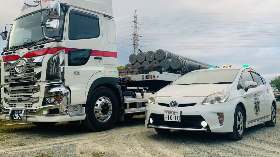 電柱を積載したトラックの隣に待機する先導車。橋村運送の先導車事業部の車両